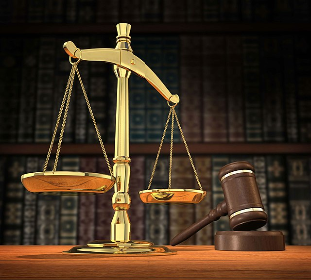 The scales of justice and a judge's gavel on an office desk with a bookshelf in the background.